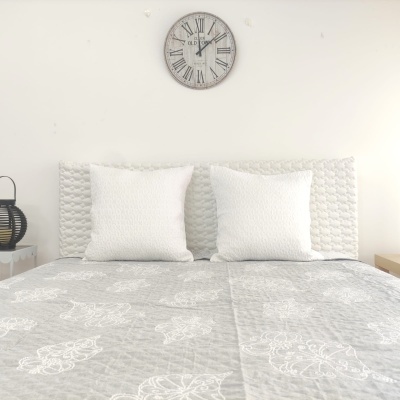 Cama com edredão cinzento e almofadas brancas, relógio de parede com números romanos