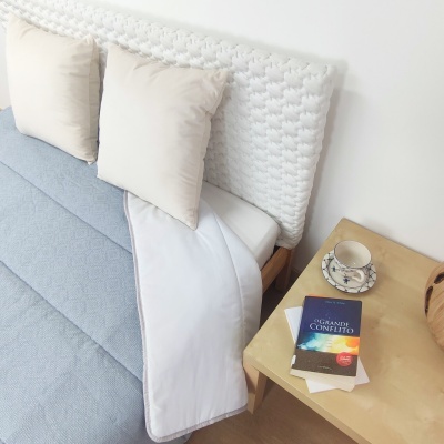 Cama com colcha azul clara, almofadas bege, cabeceira branca trançada, mesa lateral com livro, xícara e prato branco