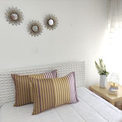 Almofadas riscas castanho lilás creme em cama com cabeceira branca, mesa de madeira com vaso e candeeiro