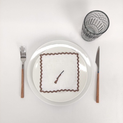 Mesa posta com prato branco, guardanapo branco com borda castanha e desenho de espingarda, garfo e faca com cabo em madeira, copo com padrão geométrico preto