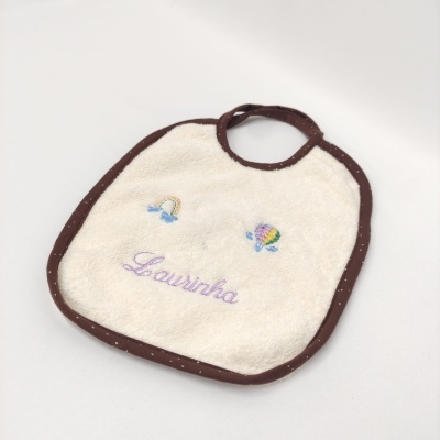 Babete creme com bordas castanhas, bordados coloridos e nome 'Laurinha' em lilás