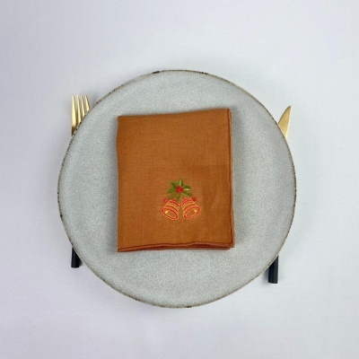 Guardanapo castanho com bordado de sinos sobre prato cinzento e talheres dourados num fundo branco