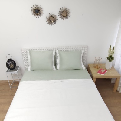 Cama com roupa de cama verde claro e branca, cabeceira trançada branca, mesas laterais com decoração, parede branca com espelhos redondos