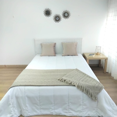 Cama com roupa de cama branca e manta cinza, duas almofadas beges, cabeceira branca entrelaçada, mesa de cabeceira em madeira clara com candeeiro e livro, parede branca com decorações circulares pretas.
