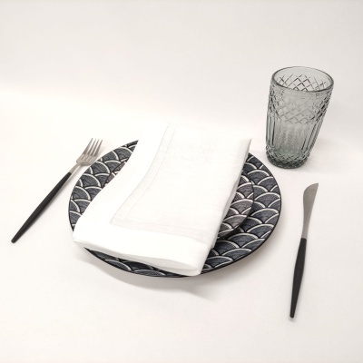 Conjunto de jantar com prato azul e branco, guardanapo branco, talheres pretos e copo de vidro cinza