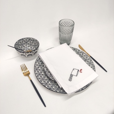 Conjunto de pratos e tigela com padrão floral azul e branco, talheres pretos e dourados, copo de vidro texturado e guardanapo branco com desenho de machado.