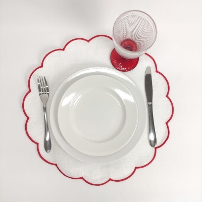 Conjunto de mesa com prato branco, talheres metálicos, copo de vidro com base vermelha e individual branco com borda vermelha em pétalas.