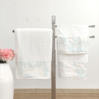 Conjunto de toalhas de banho brancas com detalhe azul pendurado em suporte metálico junto a vaso com flor