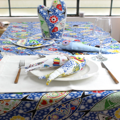 Mesa posta com toalha e acessórios de cozinha com padrão colorido de peixes e flores e três peixes de tecido no prato