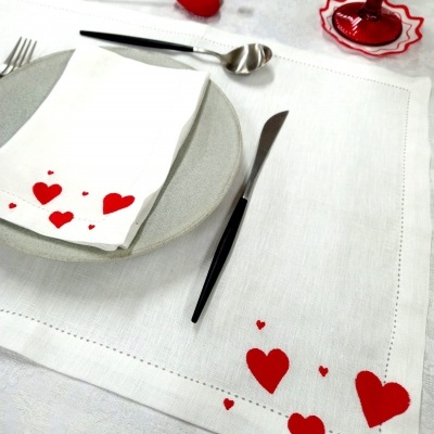Jogo americano e guardanapo branco com corações vermelhos bordados e talheres num ambiente romântico