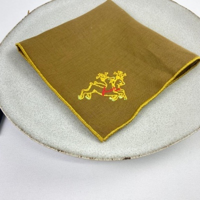 Guardanapo castanho com bordado amarelo e vermelho de dois cervos em prato cinzento