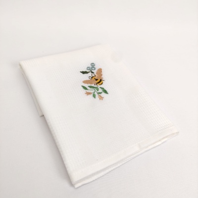 Toalha branca com bordado de abelha e flores sobre fundo branco.