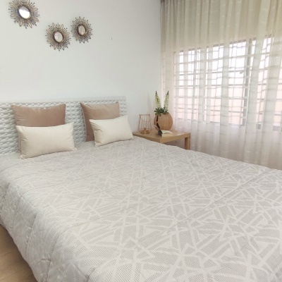 Cama de casal com cabeceira acolchoada cinza claro, roupa de cama bege com padrão geométrico, almofadas castanho e bege e mesa de cabeceira com vaso