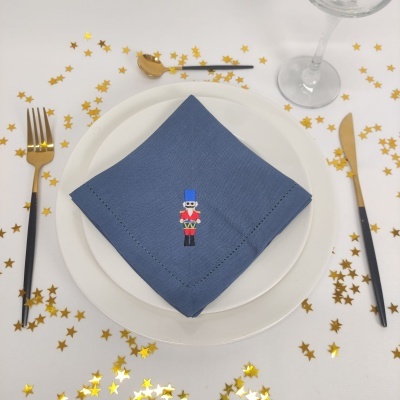 Mesa posta com prato branco, guardanapo azul com soldado quebra-nozes bordado, talheres dourados e pretos, estrelas douradas na toalha branca