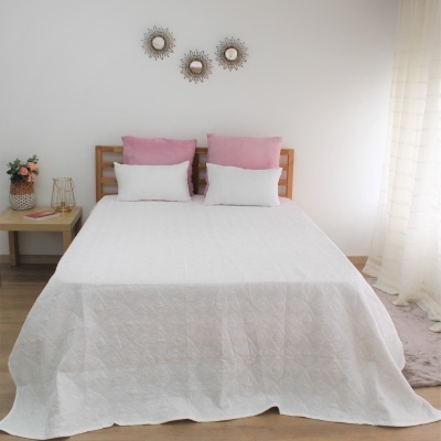 Cama de casal com colcha branca e almofadas cor-de-rosa e brancas, mesa de cabeceira com flores e candeeiro, parede branca com decorações circulares