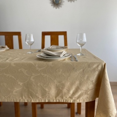 Mesa posta com toalha bege padrão floral, pratos brancos, copos de vinho, talheres e cadeiras de madeira.