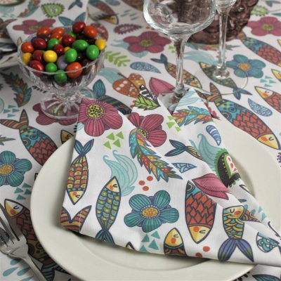Toalha de mesa e guardanapo coloridos com padrão de peixes e flores, prato branco e taças.