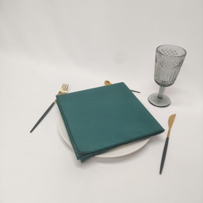 Conjunto de guardanapo verde sobre prato branco com talheres pretos e dourados e copo de vidro cinza