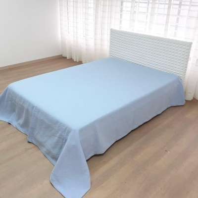 Cama com cabeceira branca acolchoada e colcha azul clara num quarto com chão em madeira clara