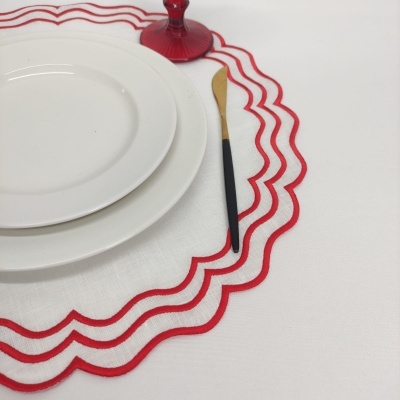 Conjunto de prato branco e marcador de mesa branco com bordado vermelho e faca com cabo dourado ao lado de copo vermelho