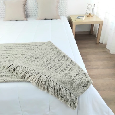 Cama com colcha branca e manta tricotada bege com franjas, almofadas bege, mesa de cabeceira em madeira clara com livro e candeeiro