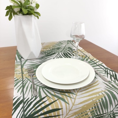Conjunto de loiça branca com talheres e copo sobre toalha de mesa com padrão folhas e vaso branco com planta