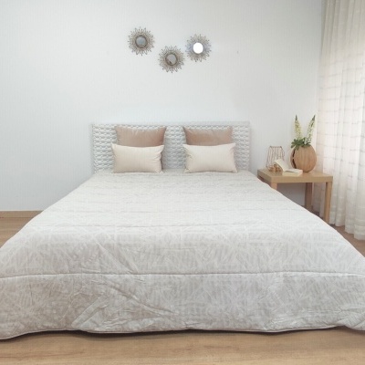 Cama de casal com roupa de cama cinza clara e almofadas bege e castanho, cabeceira branca trançada e mesa de cabeceira em madeira clara