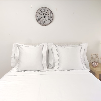 Cama com roupa de cama branca, almofadas, relógio de parede e mesa de cabeceira com abajur e relógio rosa