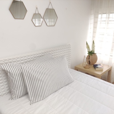 Cama branca com almofadas riscas, mesa de cabeceira com jarro e flores, espelhos geométricos na parede