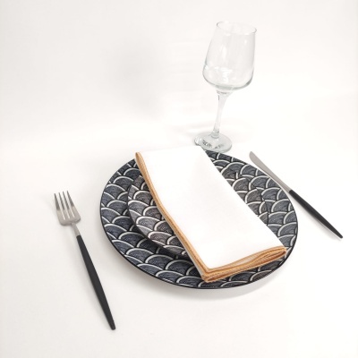 Conjunto de loiça com prato padrão geométrico azul e branco, guardanapo branco com borda bege, talheres com cabos pretos e copo de vidro