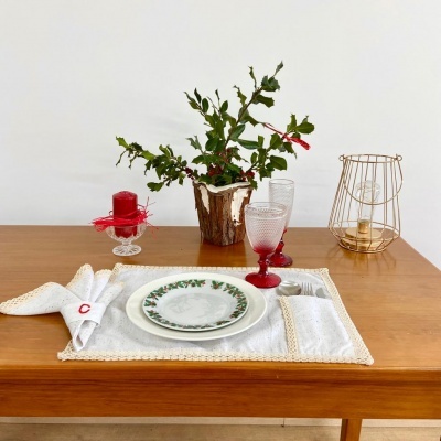 Mesa posta com prato decorativo, copos de vidro, vela vermelha, vaso com planta e suporte metálico para vela