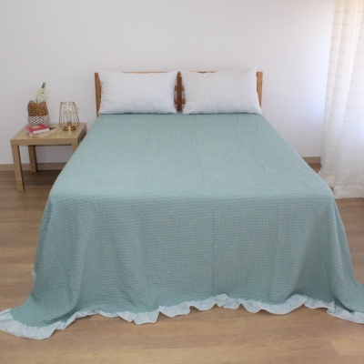 Cama de casal com colcha verde água e almofadas brancas em quarto com mesa de cabeceira e cortina branca