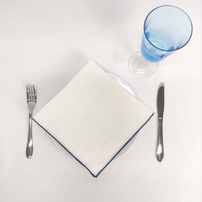 Talheres metálicos, guardanapo branco com bainha azul, prato branco e copo azul transparente num fundo branco