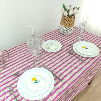 Mesa posta com toalha às riscas rosa e branca, pratos com guardanapos de flores amarelas, ovos de codorna, jarro de vidro e vaso trançado com flores brancas