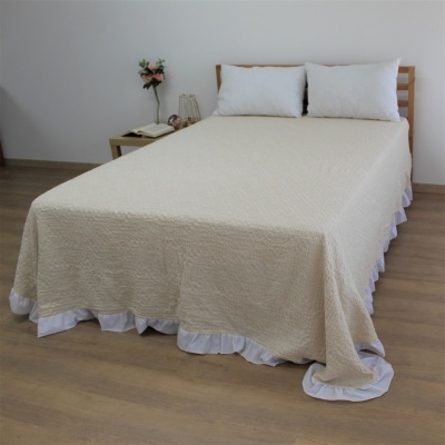 Cama com colcha bege texturizada e borda branca, duas almofadas brancas, mesa de cabeceira com flores, livro e candeeiro