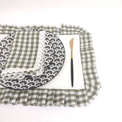 Prato branco com padrão floral preto, guardanapo xadrez verde com renda branca, individual branco com borda xadrez e faca com cabo preto e lâmina dourada