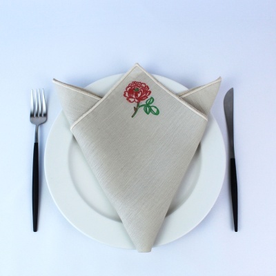 Guardanapo bege bordado com flor vermelha e folha verde sobre prato branco com talheres pretos.