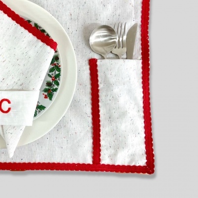 Conjunto de mesa branco com bordas vermelhas, guardanapo com letra C, prato com padrão floral, talheres no porta-talheres