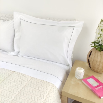 Almofada branca com borda preta sobre cama com colcha bege, ao lado mesa com vela, livro e vaso de flores