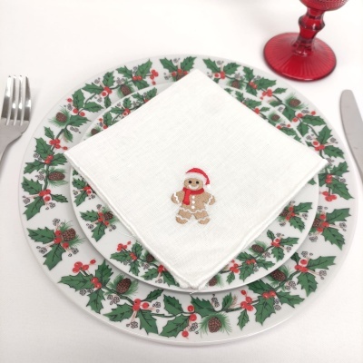 Pratos decorativos com padrão de folhas de azevinho e guardanapo branco bordado com boneco de gengibre num cenário de mesa de Natal