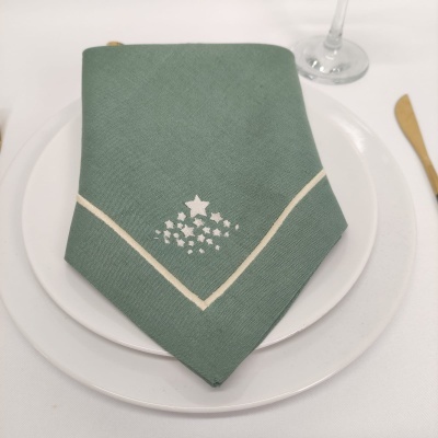 Guardanapo verde com desenho branco de árvore de Natal sobre prato branco em mesa com toalha branca.