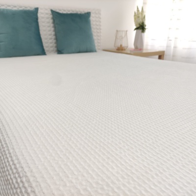 Colcha branca texturizada com almofadas azul-esverdeadas e mesa de cabeceira clara