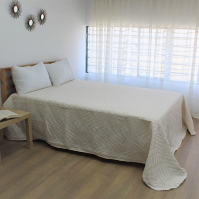 Cama de casal com colcha branca e almofadas, mesa lateral com livro, cortinas brancas transparentes
