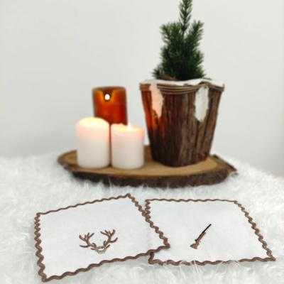 Dois porta-copos quadrados brancos com borda castanha e desenhos, velas e vaso decorativo com planta verde ao fundo