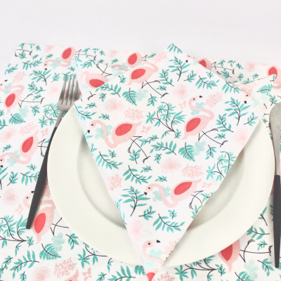 Guardanapo e toalha de mesa com padrão de pássaros cor-de-rosa e ramos verdes, prato branco, garfo e faca cinzentos.