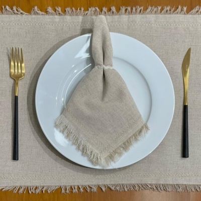 Conjunto de mesa com prato branco, guardanapo bege com franjas, garfo e faca dourados com cabos pretos sobre mesa de madeira
