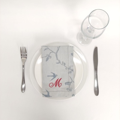 Conjunto de mesa com prato branco, guardanapo cinza com padrões, talheres metálicos e taça de vidro.