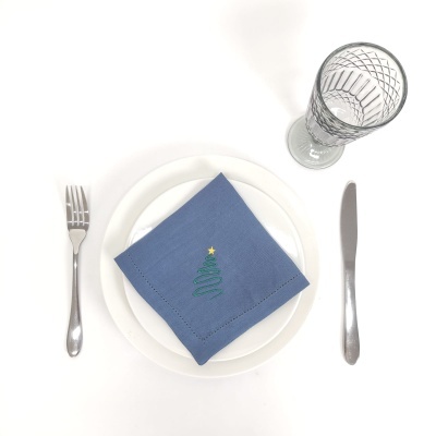 Mesa posta com prato branco, guardanapo azul com bordado de árvore de Natal, talheres de metal e copo de vidro