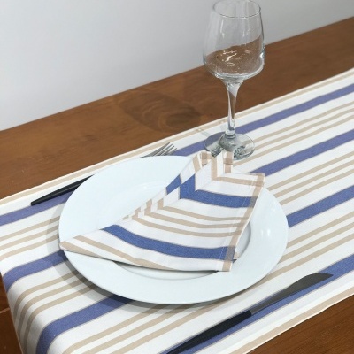 Mesa posta com jogo americano e guardanapo às riscas azuis e bege, prato branco, talheres e copo de vidro sobre mesa de madeira