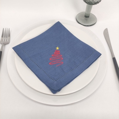 Guardanapo azul com bordado de árvore de Natal sobre prato branco com talheres e copo